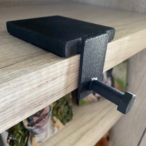 May include: A black metal shelf bracket with a textured finish. The bracket has a flat top section and a vertical support with a bolt-like projection. The design is simple and functional, suitable for various shelving needs.