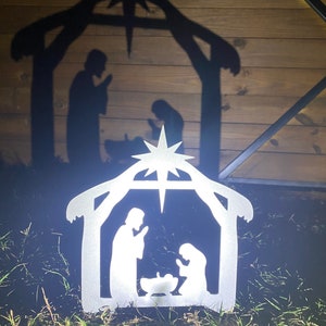 May include: A silver metal nativity scene silhouette with a star on top. The silhouette depicts Mary, Joseph, and baby Jesus in a manger. The scene is illuminated, casting a shadow of the same image on a wooden background.