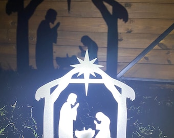 May include: A silver metal nativity scene silhouette with a star on top. The silhouette depicts Mary, Joseph, and baby Jesus in a manger. The scene is illuminated, casting a shadow of the same image on a wooden background.