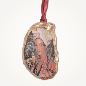 May include: An oyster shell Christmas ornament with a gold border and a claret ribbon. The image shows a woman in a turban and pink robe, with a snake, skull, and candle. The background is black.