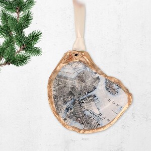 May include: An oyster shell Christmas ornament with a gold-painted edge. The ornament features a map of Boston, Massachusetts, with a cream-colored ribbon for hanging. A sprig of evergreen is in the background.