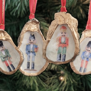 Puede incluir: Cuatro adornos navideños hechos de conchas de ostra, cada uno con un soldado cascanueces pintado. Las conchas tienen un borde dorado y están suspendidas por cintas rojas, sobre un fondo de ramas de pino.