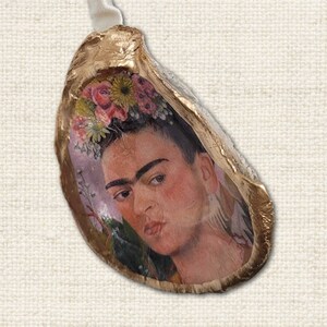 Può includere: Ornamento a conchiglia con un ritratto di Frida Kahlo. L'opera d'arte è incastonata nel guscio, che ha un bordo dorato. Un nastro bianco è attaccato per appenderlo. Il ritratto mostra Kahlo con una corona floreale.