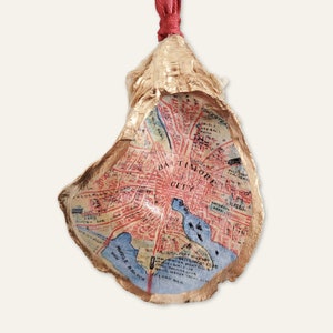 May include: An oyster shell Christmas ornament, gold-painted on the outside, featuring a map of Baltimore City inside. The map is in red and blue, with 'Baltimore City' visible. A red ribbon is attached for hanging.