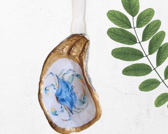 May include: An oyster shell ornament with a gold-painted rim and a watercolor crab design in shades of blue and yellow. The shell is suspended by a white ribbon, perfect for coastal decor. The background is white.