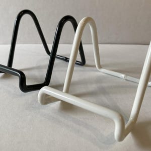 May include: Two metal display stands, one black and one white, are side-by-side. Each stand has a curved top and a horizontal base, designed to hold items upright. The stands are made of a smooth, possibly painted, metal.