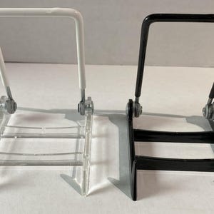 May include: Two display stands, one clear and one black, are side-by-side. Each stand has a rectangular base, a hinged support, and a top bar. The clear stand has a white top bar, while the black stand has a black top bar.
