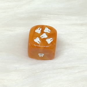 May include: A brown dice with white teeth printed on each side. The dice is made of a translucent material and has a slightly marbled appearance.