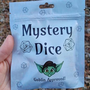 May include: A silver pouch with a zipper closure, featuring the text "Mystery Dice" and a cartoon goblin with the text "Goblin Approved!" printed on it. The pouch is decorated with multiple images of dice with question marks on them.