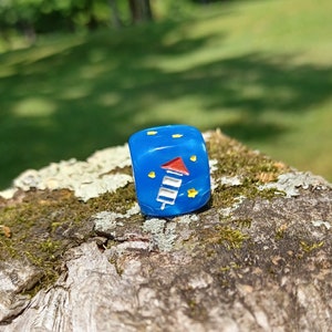 May include: A blue dice with white and red stars and a firework design. The dice is sitting on a piece of bark.
