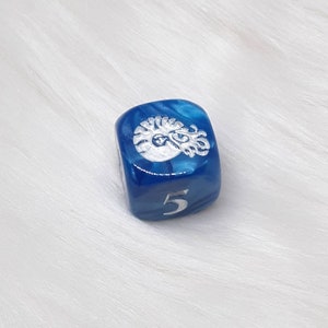 May include: A blue and white marbled six-sided die with a silver-colored design on one face and the number 5 on another. The die is set against a white, fluffy background.