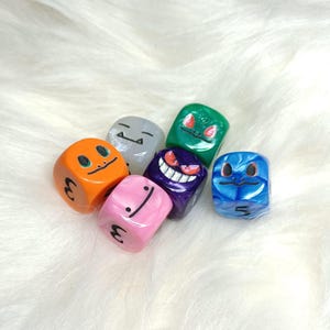 May include: Six colorful dice with different cartoon faces. The dice are made of plastic and have a glossy finish. The faces include a ghost, a green creature, a blue creature, a pink creature, an orange creature, and a white creature.