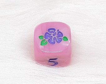 May include: Pink dice with a purple flower and green leaves on one side and the number 5 on the opposite side.
