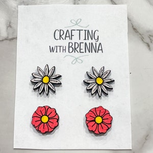 Puede incluir: Un conjunto de cuatro pendientes de madera con forma de flor. Dos pendientes tienen margaritas blancas con centros amarillos, y dos pendientes tienen flores rosas con centros amarillos.