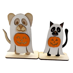Puede incluir: Dos decoraciones de Halloween de madera, un perro y un gato, vestidos de fantasmas y sosteniendo calabazas.