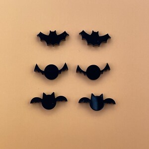 May include: Six black bat-shaped acrylic ornaments. Two bats have a solid shape, two bats have a circle cut out in the center, and two bats have a circle cut out in the center with a small bat shape inside the circle.