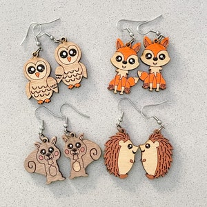 Könnte beinhalten: Satz mit sechs Holzohrringen mit Tiermotiven. Die Ohrringe sind aus Naturholz gefertigt und haben eine hellbraune Farbe. Die Designs umfassen zwei Eulen, zwei Füchse und zwei Igel.