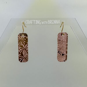 May include: A pair of rose gold earrings with a floral design. The earrings are rectangular in shape and have a gold hook closure.