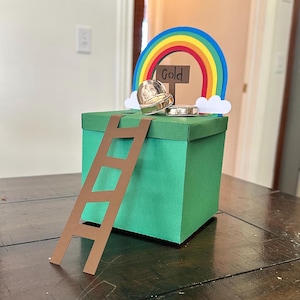 Puede incluir: Una caja verde con una escalera marrón. Un arcoíris y un letrero que dice "Gold" están encima de la caja, junto con una moneda de oro y una pequeña olla de oro. La caja está sobre una mesa de madera oscura.