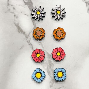 Puede incluir: Seis pares de pendientes de madera con forma de flor. Los pendientes son de varios colores, incluyendo blanco, naranja, rosa y azul. Cada par presenta un dise&ntilde;o de flor diferente.