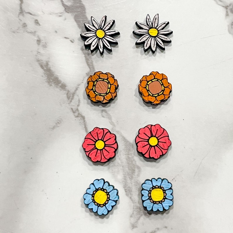 Puede incluir: Seis pares de pendientes de madera con forma de flor. Los pendientes son de varios colores, incluyendo blanco, naranja, rosa y azul. Cada par presenta un dise&ntilde;o de flor diferente.