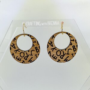 May include: A pair of wooden earrings with a gold hook. The earrings are round and have a black and white pattern of letters.
