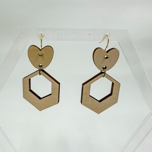 May include: A pair of wooden earrings with a geometric hexagon shape and a heart shape on top. The earrings are a light brown color and have gold hooks.