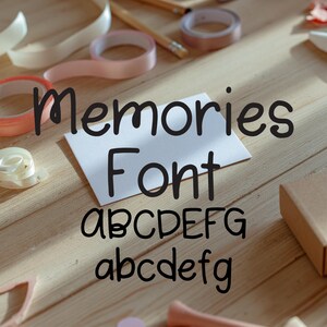 May include: A wooden surface with stationery supplies and the text "Memories Font". The text is displayed on a white card, with the alphabet in uppercase and lowercase. Rolls of tape and ribbon are scattered around the card.