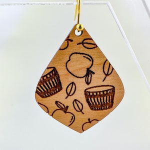 May include: A pair of wooden dangle earrings with a brown wood background and a pattern of apples, leaves, and baskets. The earrings have gold hooks.