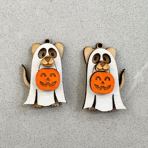 Puede incluir: Dos adornos de madera en forma de fantasma blancos con detalles marrones. Cada fantasma sostiene una pequeña calabaza naranja con una cara de calabaza.