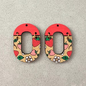 May include: A pair of wooden earrings with a red top and an oval shape. The oval section features a strawberry pattern with red strawberries, green leaves, and white flowers. Gold-colored hooks are attached.