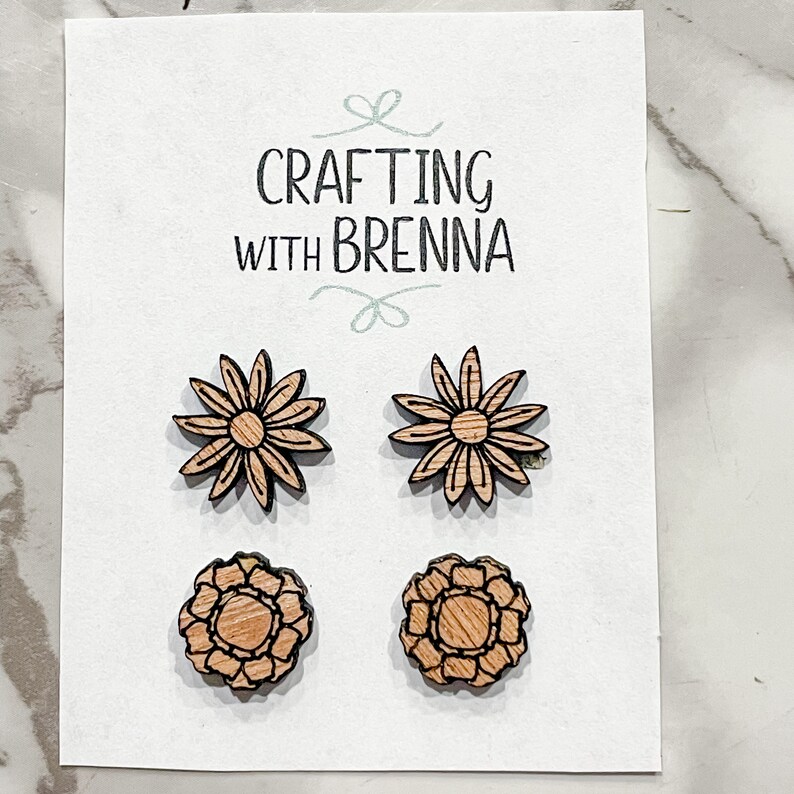 Puede incluir: Cuatro pendientes de madera con forma de flor, dos con un dise&ntilde;o de margarita y dos con un dise&ntilde;o de flor m&aacute;s intrincado. Los pendientes se muestran en una tarjeta blanca con el texto "CRAFTING WITH BRENNA".