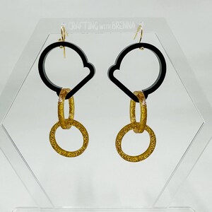 Può includere: Un paio di orecchini neri e dorati con un design geometrico. Gli orecchini presentano un grande cerchio nero con un cerchio più piccolo tagliato, seguito da due anelli dorati.