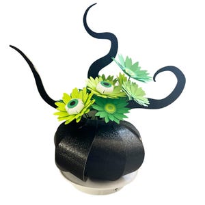 May include: A Halloween-themed centrepiece featuring a black pumpkin-shaped vase with green and yellow paper flowers. The flowers have white and green eyeball centres. Black, curved accents rise above the flowers, creating a spooky, decorative arrangement.