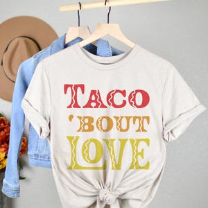 以下が含まれることがあります： ベージュのTシャツに「Taco 'Bout Love」という文字が赤、黄、オレンジでプリントされています。文字はレトロなフォントスタイルです。