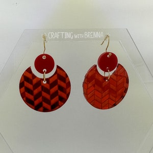 May include: A pair of red and gold geometric earrings with a chevron pattern. The earrings are displayed on a white acrylic stand.