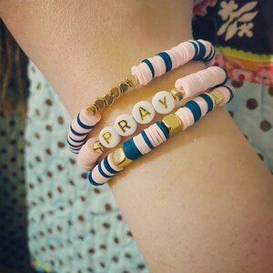 Può includere: Un set di tre braccialetti con perline rosa e blu. Il braccialetto centrale ha perline bianche con la parola "PRAY" scritta in lettere dorate.