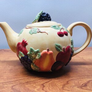 May include: A ceramic teapot with a cream-colored base and a colorful fruit design. The teapot features a variety of fruits, including apples, pears, grapes, and cherries, all painted in realistic detail.