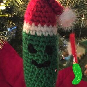 K&ouml;nnte beinhalten: Ein gr&uuml;ner, gestrickter Gurken-Anh&auml;nger mit einer rot-wei&szlig;en, gestrickten Weihnachtsmannm&uuml;tze. Die Gurke hat ein schwarzes, l&auml;chelndes Gesicht.
