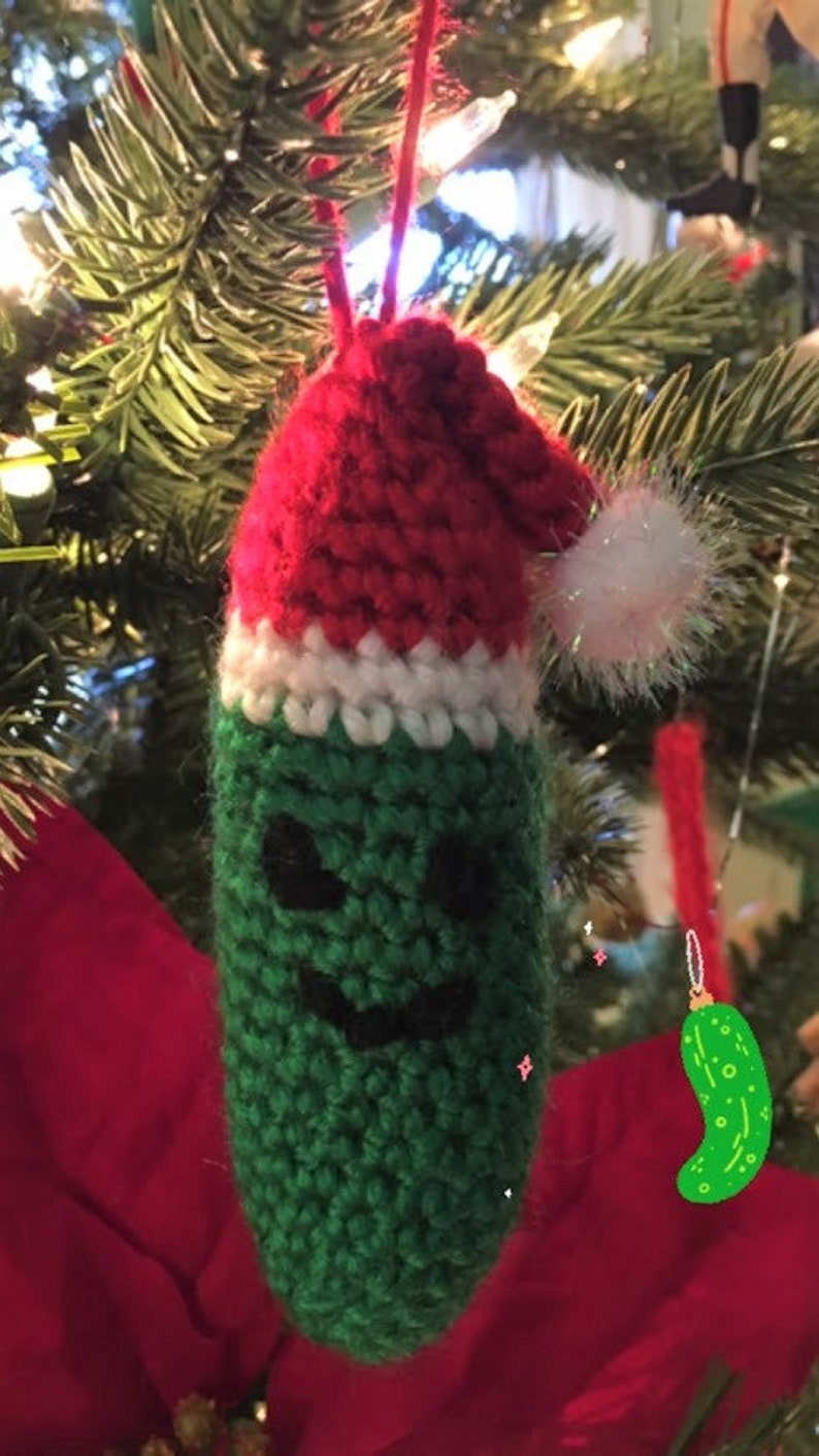 K&ouml;nnte beinhalten: Ein gr&uuml;ner, gestrickter Gurken-Anh&auml;nger mit einer rot-wei&szlig;en, gestrickten Weihnachtsmannm&uuml;tze. Die Gurke hat ein schwarzes, l&auml;chelndes Gesicht.