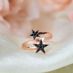 May include: A rose gold ring with two black star-shaped settings. The ring is designed to be worn on the middle finger.