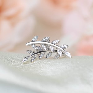 May include: A silver-colored ear cuff in the shape of a stylized leaf branch. The branch curves gracefully, adorned with small, sparkling clear gemstones. The jewelry piece rests on a soft, white fabric, with a blurred pink background.