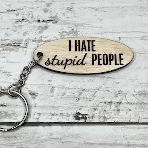 May include: Oval wooden keychain with the phrase "I HATE stupid PEOPLE" engraved in black. The keychain has a silver chain and a keyring. The background is a white and gray wood grain.