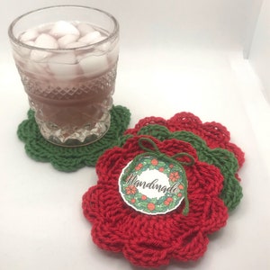 May include: Set of three crocheted coasters in red and green. The coasters are shaped like flowers and have a scalloped edge. A tag with the word "Handmade" is attached to the coasters.