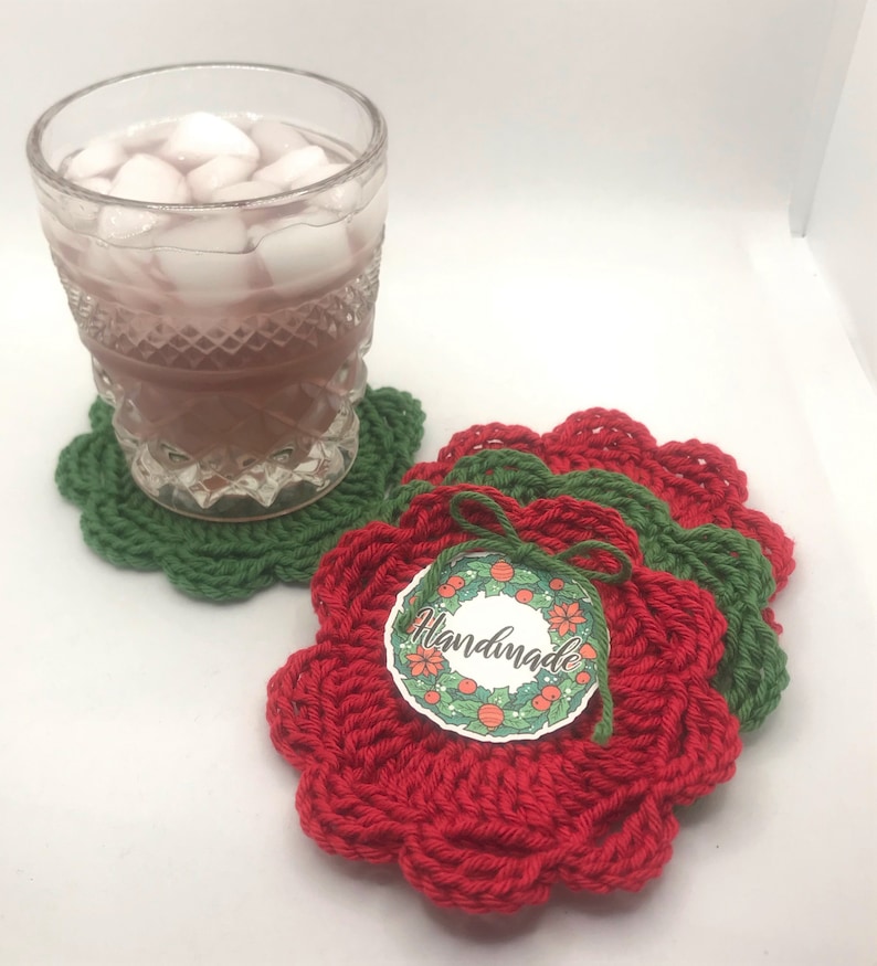 May include: Set of three crocheted coasters in red and green. The coasters are shaped like flowers and have a scalloped edge. A tag with the word "Handmade" is attached to the coasters.