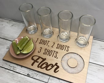 Può includere: Porta bicchieri da shot in legno con quattro bicchieri da shot, un piccolo piatto rosa con spicchi di lime e un bordo di sale. Il supporto ha il testo "1 shot, 2 shots, 3 shots, Floor" inciso su di esso.