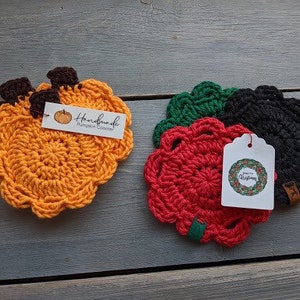 May include: Three crocheted coasters, one orange, one red, and one black. The orange coaster is shaped like a pumpkin and has a tag that says "Handmade Pumpkin Coaster". The red and black coasters have a tag that says "Merry Christmas".
