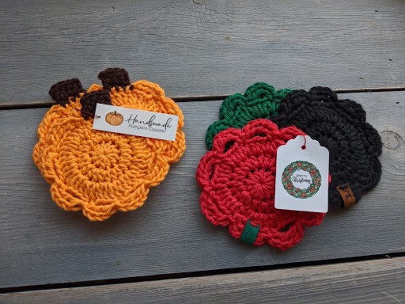 May include: Three crocheted coasters, one orange, one red, and one black. The orange coaster is shaped like a pumpkin and has a tag that says "Handmade Pumpkin Coaster". The red and black coasters have a tag that says "Merry Christmas".