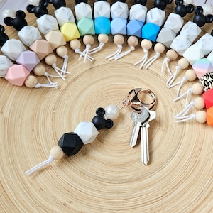 May include: A set of colorful silicone and wooden bead keychains with a Mickey Mouse head charm. The keychains are arranged in a semi-circle on a wooden surface. Each keychain has a different color and pattern, including marble, pastel, and solid colors. The keychains are attached to a gold-colored key ring with two keys.