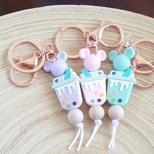 May include: Three keychains featuring colorful boba tea cup charms with mouse ear accents. Each keychain has a rose gold ring and clasp. The boba tea charms are pastel purple, pink, and teal, with wooden bead accents.